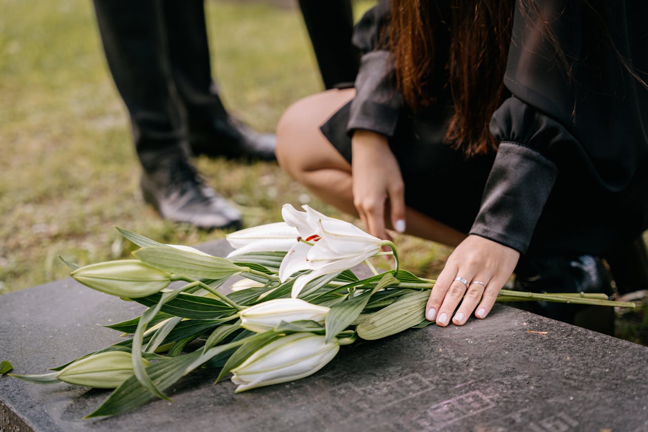 Services A woman in black attire mourns beside a grave, placing white lilies on the headstone in a cemetery setting.